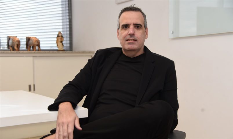 Foto de Marcelo Cohen sentado sobre mesa com perna cruzada, vestindo roupas pretas.