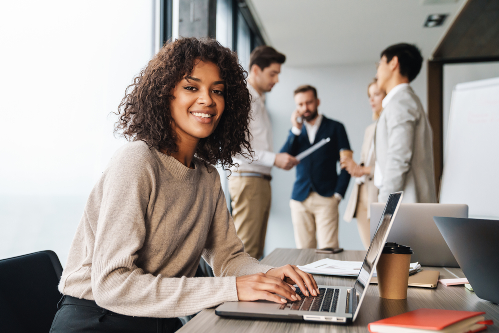 Mulher morena sorrindo para a câmera e utilizando um computador. Grupo de pessoas atrás vestidas formalmente conversando. Ambiente limpo e claro.